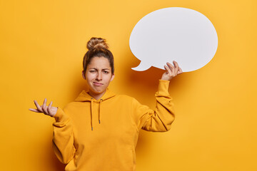 Hesitant puzzled young woman with hair bun shrugs shoulders holds blank communication bubble for your advertising content doesnt know what to write here isolated over vivid yellow background - obrazy, fototapety, plakaty