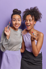 Vertical shot of two women stand closely to each other apply red lipstick do makeup wears...