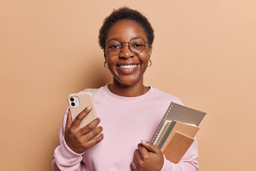 Positive dark skinned African girl with toothy smile holds mobile phone and notepads studies at...