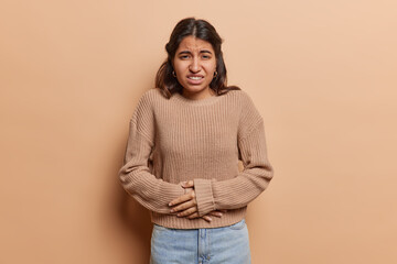 Unhappy sick Iranian woman keeps both hands on stomach suffers from pain or menstruation cramps frowns face wears knitted jumper and jeans isolated over brown background. Health problems concept