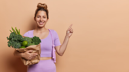 Health and sport. Indoor photo of young happy fit smiling European lady standing on left isolated on beige background with big paper bag of vegetables wearing purple tracksuit pointing at blank space