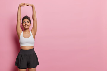 Horizontal shot of Iranian woman keeps arms raised up dressed in white cropped top and shorts being in good physical shape isolated over pink background copy space for your promotional content