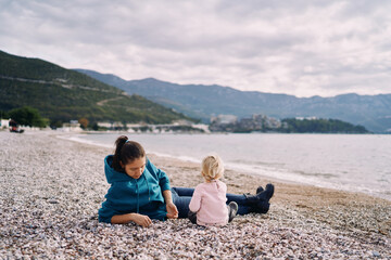 Mom with a little girl are sitting on the beach by the sea and playing with pebbles