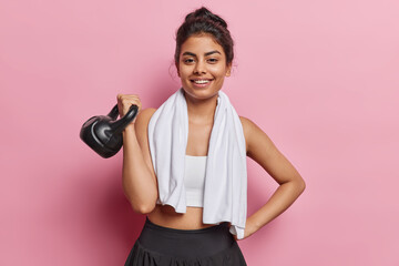 Horizontal shot of cheerful motivated Iranian woman lifts weight and smiles broadly poses with towel around neck dressed in activewear poses against pink background. Sport and exercising concept