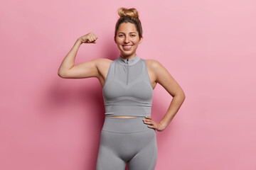 Positive European woman flexing biceps and smiles happily dressed in sports clothing smiles gladfully maintaines active lifestyle for good health poses over pink background feels proud of achievements