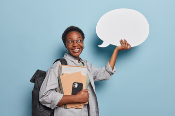 Horizontal shot of curly haired teenage girl poses with notepads smartphone and white blank communication bubble for your text poses with rucksack isolated over blue background. Studying concept