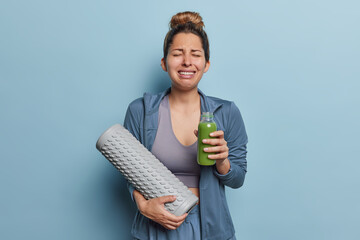 Upset doleful sportswoman holds bottle of vegetable juice and foam roller has frustrated expression...
