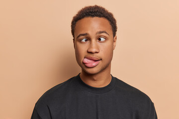 Studio close up of young funny African american man with short curly hair wearing black t shirt standing in centre isolated on beige background keeping hands down showing tongue making grimace - obrazy, fototapety, plakaty