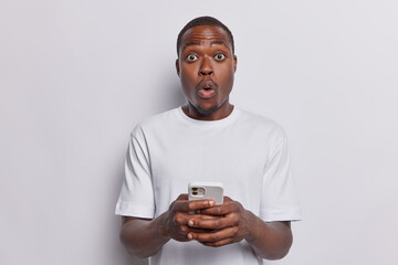 Waist up shot of stunned dark skinned adult man holds mobile phone in hands shocked being bullied online reads unexpected message dressed in casual basic t shirt isolated over white background