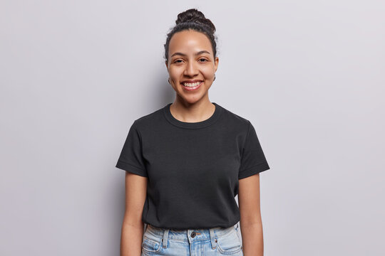 Waist up shot of cheerful Latin woman with combed hair and pleasant smile on face dressed in casual black t shirt annd jeans concentrated at camera isolated on white background. Happy face expressions