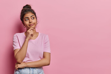 Horizontal shot of pretty young Iranian woman keeps hand on chin concentrated aside thinks about something dressed in casual t shirt and jeans isolated over pink background. Hmm let me think