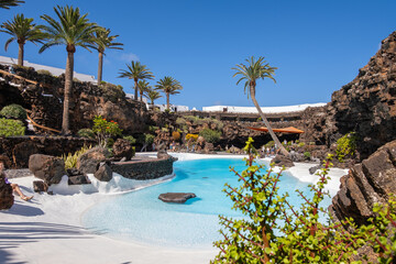 Pool of blue waters and palemeeras, outside the cave of Los Jameos del Agua. Light at the end of the cave. Sky with big white clouds. Lanzarote, Canary Islands, Spain.
