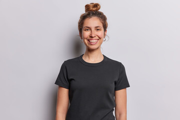 Waist up shot of pretty cheerful European woman with glad smile on face looks directly at camera dressed in casual black t shirt expresses positive emotions isolated over white studio background
