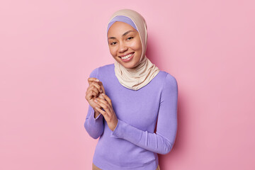 Indoor shot of positive Muslim wears hijab and purple turtleneck smiles gently keeps hands together...