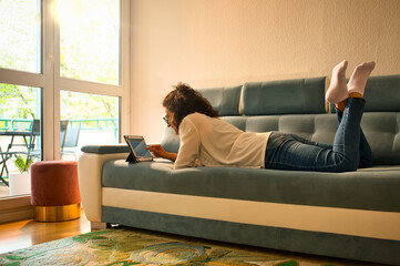 Young woman surfs the net on a tablet lying on the couch and smiling in warm sun light with sun peeking through the window