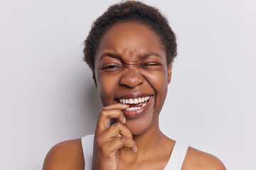 Studio shot of dark skinned adult woman with curly hair bites finger and smirks face shows white teeth poses against white background winks eye has happy expression. People and emotions concept - obrazy, fototapety, plakaty