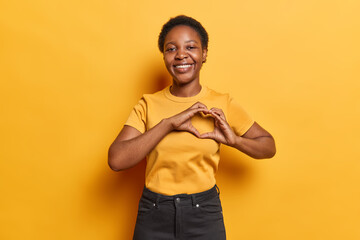 Horizontal shot of African woman shows heart love sign smiles gladfully dressed in casual t shirt...