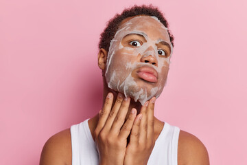 Skin care procedures. Dark skinned young African man applies beauty sheet mask on face for reducing...