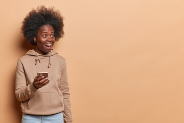 Fototapeta na wymiar Happy surprised dark skinned woman with curly bushy hair enjoys browsing social media reacts to something amazing wears casual sweatshirt and jeans isolated over brown background copy space aside