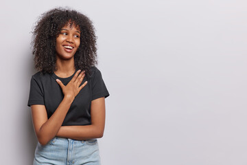 Fototapeta na wymiar Horizontal shot of cheerful African woman with curly hair keeps hand on chest smiles gladfully and focused aside wears black t shirt jeans isolated over white background copy space for your promotion