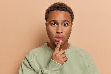 People and emotions concept. Stunned dark skinned handsome man keeps finger on lips stares...