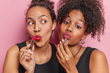 Horizontal shot of two friendly women apply lipstick on lips does makeup at home get prepare for...