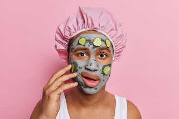 Photo of young man examines his skin applies beauty clay mask with cucumber slices on face for rejuvenation and skin regeneration wears bath hat and white t shirt isolated over pink studio wall