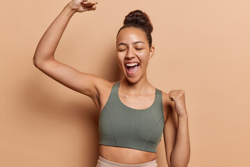 Horizontal shot of dark haired sporty woman makes winner gesture celebrates success dressed in sport bra feels jubilant exclaims loudly isolated over brown background. Yes finally I achieved myy goals