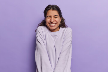 Waist up shot of positive Iranian woman with dark hair smiles happily keeps eyes closed recalls something funny dressed casually isolated over purple background. Positive human emotions concept