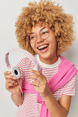 Vertical shot of cheerful carefree woman with curly hair holds stereo headphones smiles broadly wears transparent eyeglasses striped t shirt and jumper over shoulders enjoys listening favorite music