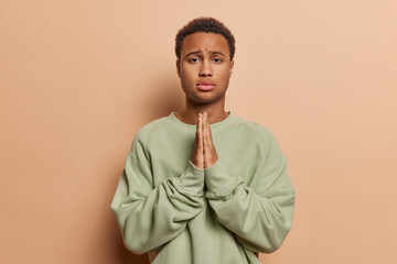 Waist up shot of African man with dark curly hair keeps palms pressed together asks for advice...