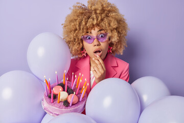 Fototapeta na wymiar People reactions and celebration concept. Stunned curly haired woman stares bugged eyes at camera keeps mouth opened poses with tasty festive cake poses among balloons against purple background