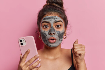 Horizontal shot of stunned woman applies facial clay mask on face for skin treatment and reducing...