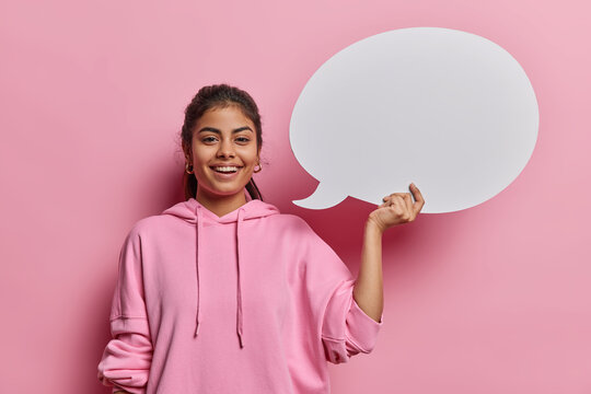 Positive lovely Iranian woman with dark hair smiles gladfully holds blank speech bubble gives promotional opportunity smiles gladfully wears casual sweatshirt poses against pink studio background.
