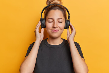 Indoor shot of pretty young woman with hair bun enjoys audio app wears black headphones on ears has...