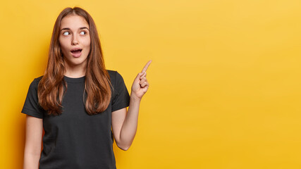 Horizontal shot of surprised long haired woman dressed in casual black t shirt has wondered curious expression points index finger on copy space for your advertisement isolated over yellow background