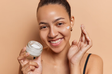 Skin hydration concept. Positive good looking Latin woman applies moisturising cream on face uses...