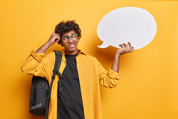 Hesitant clueless Hindu man scratches head and feels unaware thinks which words to fill on this empty speech bubble poses with rucksack on shoulder isolated over yellow background makes up his mind