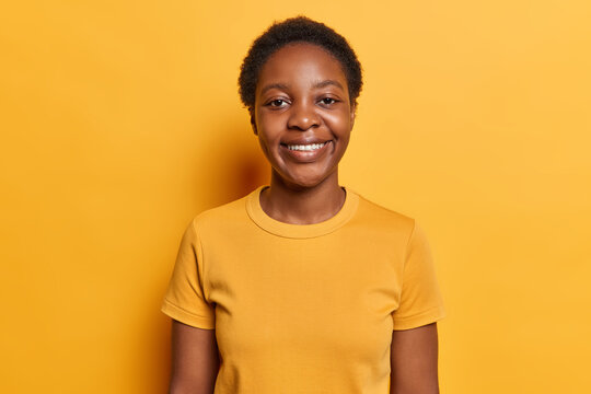 Horizontal shot of good looking millennial girl with curly hair happ smile on face expresses positive emotions dressed in casual t shirt looks gladfully at camera isolated over yellow background.