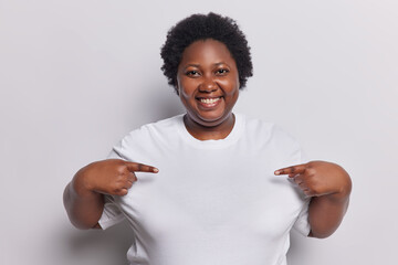 Clothing template concept. Positive overweight African woman with curly hair points at copy space of t shirt suggests to write you logo here isolated over white background. Place you test here