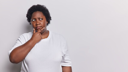 Unsure hesitant African woman with curly hair holds chin concentrated aside has doubtful expression dressed in casual clothes isolated over white background copy space for your promotional content - obrazy, fototapety, plakaty