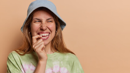 Horizontal shot of positive European woman bites finger and looks aside smiles gladfully has curious expression notices something funny wears panama and t shirt isolated over brown background