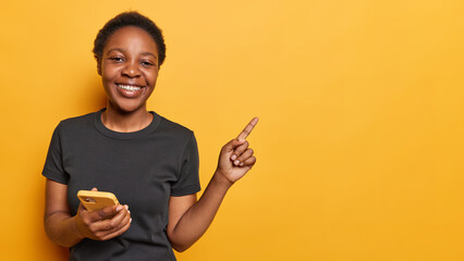 Waist up shot of positive dark skinned woman holds mobile phone pointing index finger aside showing...