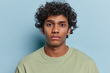 Fototapeta na wymiar Portrait of handsome curly haired young Hindu man wears basic t shirt looks directly at camera with focused expression poses against blue studio background. People and facial expressions concept