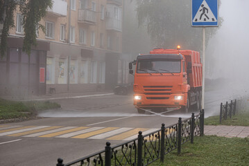 A truck washes the road with water in the early morning during fog. Cityscape.