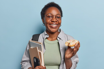 Smiling dark skinned teenage girl with short curly hair eats glazed doughnut holds notepads with smartphone has delicious snack wears spectacles and casual clothing isolated over blue background. - Powered by Adobe