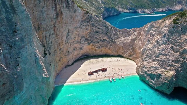 Navagio Pebble Beach between towering cliffs with a shipwreck and turquoise water. Aerial view of tourists enjoying summer vacation at the cove reached by boats, Navagio Beach with crystal blue water.