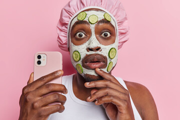 Photo of stunned dark skinned man poses with smartphone in hand applies nourishing clay mask with...
