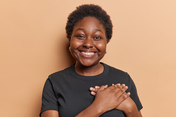 Dark skinned chubby woman smiles broadly presses hands to heart expresses gratitude expresses genuine appreciation wears casual black t shirt isolated over brown background. People and thankfulness