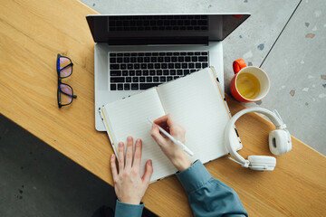 A woman takes notes in a notepad using a laptop sitting at the table, top view. Workplace with...